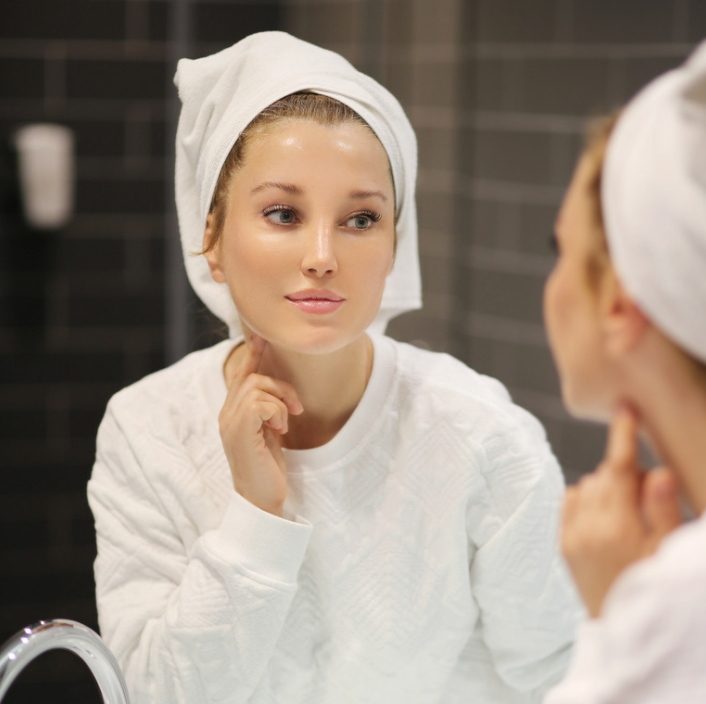 Woman applying Herbal Relief Cream in front of a mirror with a towel on her head, showcasing skincare routine.