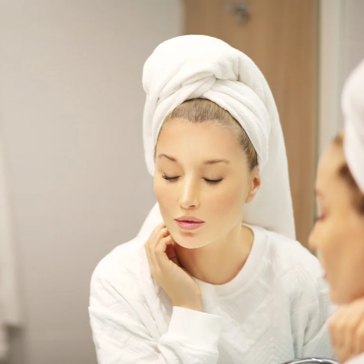 Woman with a towel on her head, showing a serene facial expression in a mirror, emphasizing self-care routine.