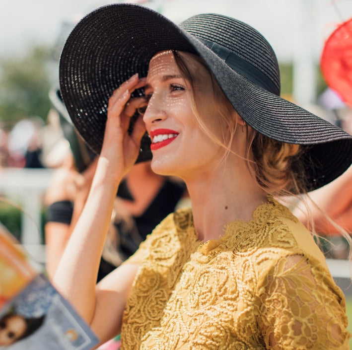 A woman in a black wide-brim hat and yellow lace dress smiling outdoors at an event.