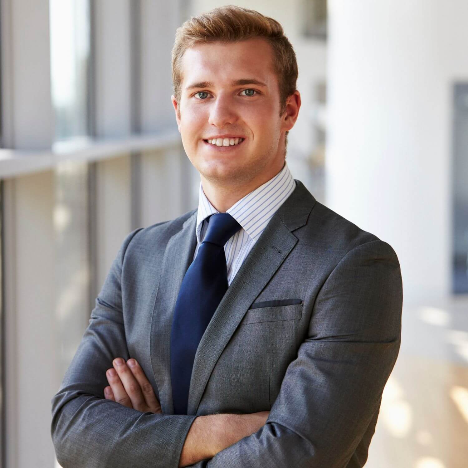 Smiling young professional in a suit and tie, standing confidently in a modern office space.